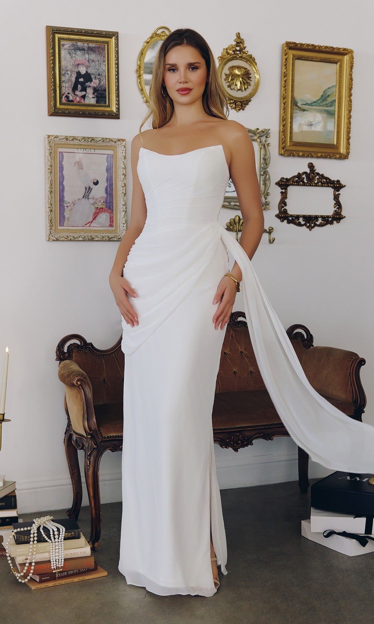A woman wears the Amoris T726W Long Off-White Formal Dress with an adjustable lace-up back, standing indoors by framed art, a vintage loveseat, and stacked books.